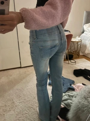 Ljusblå bootcut jeans med slitningar - Säljer ett par ljusblå bootcut jeans med snygga slitningar och klassiska fem fickor. Jeansen har normalhög midja och stängs med dragkedja och knapp. Perfekta för en avslappnad och trendig look.