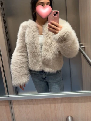 Beige fluffig pälsjacka - Supermjuk och fluffig beige pälsjacka med bred krage och långa ärmar. Jackan har en kort, boxig passform och är perfekt för att hålla värmen under kalla dagar. Snygg att styla med jeans eller klänning.