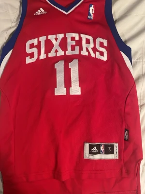 Röd Sixers basketlinne Adidas S - Snyggt rött basketlinne från Adidas med Philadelphia 76ers tryck, nummer 11 och namnet Holiday på ryggen. Linnet har vita och blå detaljer, NBA-logga och är tillverkat i ett lätt, ventilerande material perfekt för träning eller match.