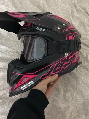 Svart och rosa crosshjälm Just1 - Säljer en svart crosshjälm från Just1 med rosa detaljer och coola grafiska mönster. Hjälmen har en stor skärm framtill och medföljande genomskinliga glasögon med svart band där det står RAVE. Perfekt för motocross eller enduro. Säljer den då den aldrig kommer till användning, en liten repa på vänster sida men inget som syns massor! Den är i storlek XS🤍