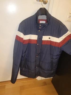 Blå pufferjacka från U.S. Polo Assn - Mörkblå pufferjacka med vita och röda ränder över bröstet från U.S. Polo Assn. Jackan har hög krage, dragkedja och tryckknappar framtill samt två fickor med dragkedja. Materialet är glansigt och quiltat för extra värme.