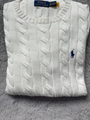 Vit kabelstickad tröja Polo Ralph Lauren - Kabelstickad vit tröja från Polo Ralph Lauren med rund halsringning och klassisk blå logga broderad på bröstet. Tröjan har ribbade muddar vid ärmslut och nederkant, vilket ger en snygg och tidlös look. Perfekt för dig som gillar stilrena plagg. Storlek XS men lite sot ri storleken sitter mer som en M 