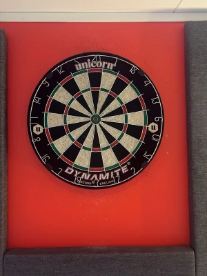 Unicorn darttavla Dynamite - Säljer en klassisk rund darttavla från Unicorn, modell Dynamite. Tavlan har svart, vit, röd och grön färg med tydliga poängmarkeringar och metalltrådar som delar fälten. Perfekt för dartspel hemma eller med vänner.