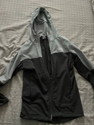 Grå och svart vindjacka The North Face - Snygg vindjacka från The North Face i grått och svart. Jackan har huva, hel dragkedja framtill och två sidofickor. Tillverkad i ett lätt och vindtätt material, perfekt för blåsiga dagar. Logotyp på bröstet och sportig passform. Använd ett par gånger men inga defekter på den 