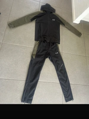  SÖKER Under Armour svart träningsset  -  SÖKER UNDER ARMOUR TRQCJSUITES Snyggt svart träningsset från Under Armour med olivgröna detaljer på ärmar och sidor. Setet består av en hoodie med dragkedja och matchande byxor med smal passform. Perfekt för gymmet eller löpning. Materialet är troligtvis polyester för bra andningsförmåga.