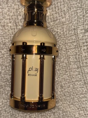 Reham parfym från Khadlaj - Lyxig parfymflaska från Khadlaj i modellen Reham. Flaskan är krämfärgad med guldfärgade detaljer och har en elegant, rundad form med dekorativa pelare. Perfekt för dig som gillar exklusiva dofter och snygg design. Ca 90% kvar 