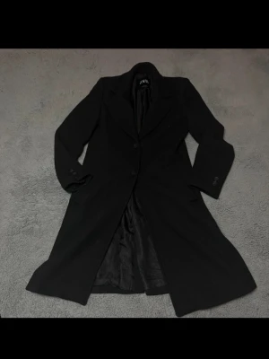 Svart lång rock från Zara - Stilren svart lång kappa från Zara med klassisk krage och två knappar framtill. Kappan har en rak passform och är helfodrad med slätt innerfoder. Perfekt för dig som vill ha en tidlös och elegant look.