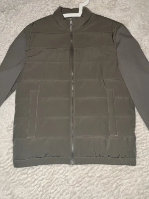 Olivgrön pufferjacka med dragkedja - Snygg o pufferjacka med raka linjer och quiltad design. Jackan har två sidofickor, hög krage och hel dragkedja framtill. Ärmarna är i matchande tyg och jackan har en minimalistisk look som passar till många stilar. Använd 2 gånger annars bara legat i garderoben. 