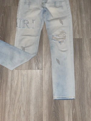 Ljusblå slitna AMIRI jeans - Säljer ett par ljusblå jeans med slitningar och tryckdetaljer på ena benet. Jeansen har raka ben och en avslappnad passform. Snygga detaljer med slitna partier och textapplikationer som ger en trendig look.