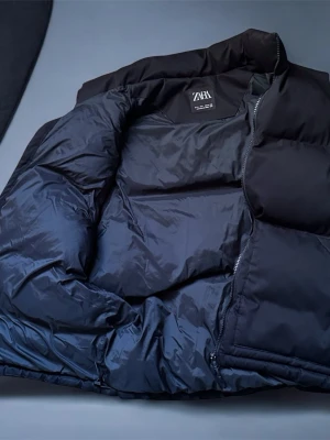 Svart dunväst från ZARA - Stilren svart dunväst från ZARA med hög krage och dragkedja framtill. Västen har två stora fickor på framsidan och är tillverkad i ett glansigt.