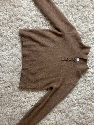 Beige stickad tröja med knappar - Mysig beige stickad tröja med långa ärmar och ribbade muddar. Tröjan har en hög krage och fyra dekorativa knappar framtill. Perfekt för lager på lager och enkel att matcha med olika outfits.