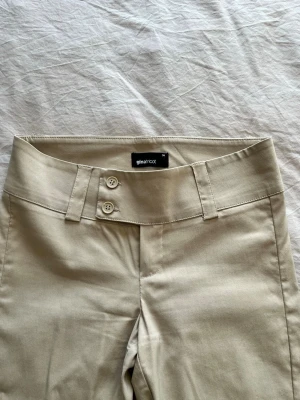 Beige chinos från Gina Tricot - Snygga beige kostymbyxor från Gina Tricot med låg midja och dubbla knappar framtill. 