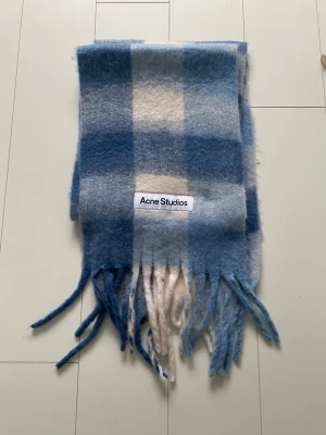Blårutig halsduk från Acne Studios - Mjuk och fluffig halsduk från Acne Studios i rutigt mönster med blå och vita toner. Halsduken har långa fransar i ändarna och en tydlig Acne Studios-logga. Perfekt accessoar för att lyfta din outfit med en trendig touch.