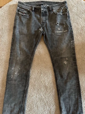 Grå slitna jeans från Diesel - Säljer ett par grå jeans från Diesel med slim fit och slitna detaljer på både fram- och baksida. Jeansen har klassisk femficksmodell, knappgylf och coola slitningar för en trendig look. Perfekta för dig som gillar streetstyle. Lagat hål imellan benen men de syns ej när dom är på!!!