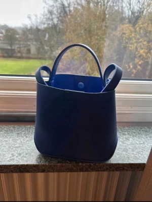 Blå handväska i konstläder -  blå handväska med rundad form och två handtag. Väskan har ett magnetlås och ett innerfack med dragkedja för småsaker. 