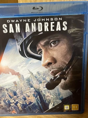San Andreas Blu-ray med Dwayne Johnson - Blu-ray-film 'San Andreas' med Dwayne Johnson i huvudrollen. Omslaget är blått och motivet visar en katastroffilm med helikopter och stadsmiljö. Filmen har åldersgräns 12 år och innehåller extramaterial som borttagna scener och gag reel.