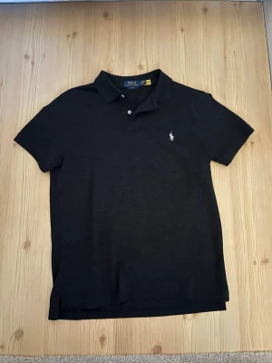 Svart pikétröja från Polo Ralph Lauren - Klassisk svart pikétröja från Polo Ralph Lauren med korta ärmar och vit broderad logga på bröstet. Tröjan har krage och knappar vid halsen. Perfekt till en avslappnad men ändå stilren look.