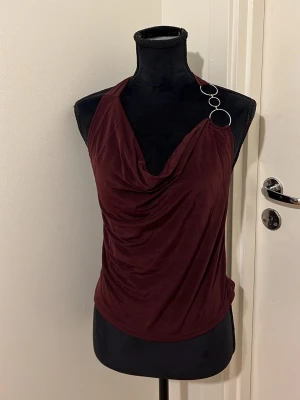 Vinröd halterneck topp med ringdetaljer - Snygg vinröd halterneck topp med draperad front och coola silverringar på ena axeln. Toppens material är mjukt och har ett lätt fall, perfekt för att skapa en trendig look. Passformen är normal och toppen är ärmlös.