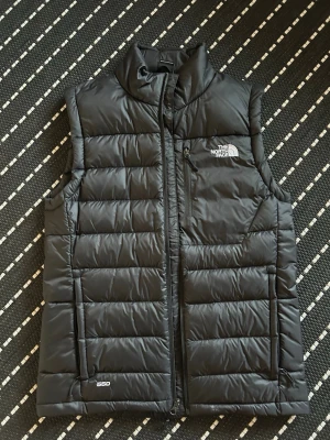 Svart dunväst från The North Face - Snygg svart dunväst från The North Face med hög krage och dragkedja framtill. Västen har en bröstficka med dragkedja, två sidofickor och diskret logga på bröstet och ryggen. Tillverkad i lätt och värmande dunmaterial, perfekt för lager på lager.