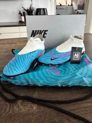 Nike Phantom GT2 Elite AG fotbollsskor - Säljer ett par Nike Phantom GT2 Elite AG fotbollsskor i blått och vitt med rosa detaljer. Skorna har strumpliknande skaft, mönstrad ovandel för bättre bollkontroll och AG-yttersula med rosa dobbar. Perfekta för konstgräs och riktigt snygg design.