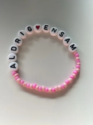 Pärlarmband med text 'Aldrig Ensam' - Sött armband med rosa pärlor, där de vita pärlorna har svarta bokstäver som bildar texten 'ALDRIG ENSAM' och ett rött hjärta. Armbandet är elastiskt och har en ungdomlig, lekfull stil.