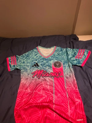 Inter Miami FC Adidas matchtröja - Säljer en färgstark Inter Miami FC matchtröja från Adidas med unikt mönster i turkos, rosa och vitt. Tröjan har korta ärmar, V-ringning och tryckta detaljer som klubbmärke, Adidas-logga och texten 'Miami' på framsidan. Tillverkad i lätt och ventilerande material.