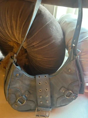 Ljusmörkblå axelväska med nitar och ringar - Säljer en ljusmörkblå axelväska i syntetmaterial med coola metalldetaljer som nitar och ringar. Väskan har en bred axelrem och ett bälte med hål och spänne framtill för extra edge. Perfekt för dig som gillar en lite tuffare stil.