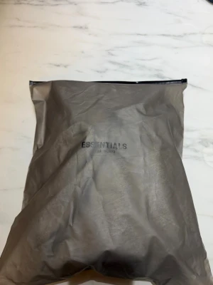 Grå Essentials Fear of God hoodie - Säljer en grå hoodie från Fear of God Essentials med stor svart logga på bröstet. Hoodien har klassisk huva och är gjord i mjukt bomullsmaterial för maximal komfort. Perfekt för dig som gillar streetwear och vill ha en stilren look.