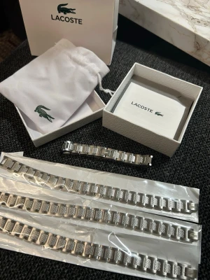Silverfärgat armband från Lacoste (justerbar) - Snyggt armband i silverfärgad metall från Lacoste med länkdesign och detaljerade mönster på varje länk. Armbandet har den klassiska Lacoste-loggan ingraverad på mittdelen (förpackningen följer inte med och säljs därför billigare) hör gärna av vid funderingar!