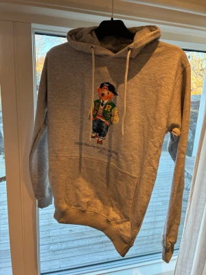 Grå hoodie med Polo Bear helt ny - Snygg grå hoodie från med den ikoniska Polo Bear-figuren på bröstet. Hoodien har huva med dragsko, magficka och ribbade muddar. Perfekt för en avslappnad och trendig stil. Materialet är mjukt och bekvämt, troligen bomullsblandning.