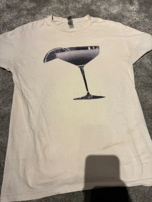 Beige t-shirt med cocktailglas print - Säljer en beige t-shirt med ett stort svart print av ett cocktailglas på framsidan. T-shirten har klassisk passform och rund hals. Perfekt för dig som gillar stilrena och grafiska motiv.