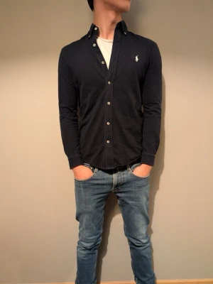 Mörkblå skjorta från Ralph Lauren - Klassisk mörkblå skjorta från Ralph Lauren med knappstängning och ikonisk logga på bröstet. Tillverkad i lätt bomullsmaterial som känns skönt mot huden. Skjortan har lång ärm och en tidlös, stilren look.