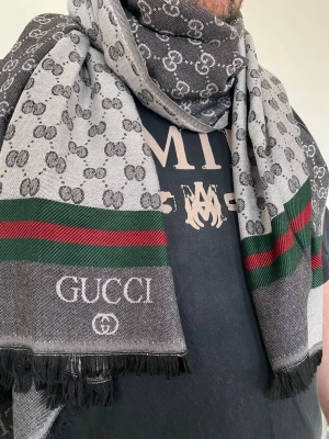 Grå halsduk från GG med logga - Snygg grå halsduk från GG med klassiskt GG-mönster, gröna och röda ränder samt vit logotyp. Halsduken har svarta fransar i kanterna och är tillverkad i ett mjukt tyg som känns lyxigt. Perfekt accessoar för att lyfta din outfit.