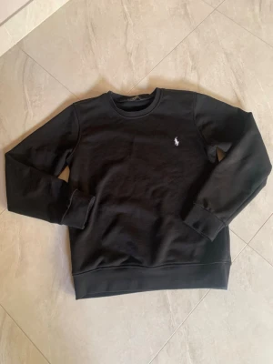 Svart sweatshirt från Ralph Lauren - Klassisk svart sweatshirt från Ralph Lauren med rund halsringning och den ikoniska vita logotypen broderad på bröstet. Tröjan har ribbade muddar vid ärmslut och nederkant. Perfekt för en clean och stilren look.