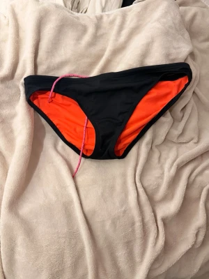 Svart träningsbikini från TYR - Säljer ett par sportiga bikini underdel från TYR i storlek XL. Yttertyget är svart och insidan är knallorange, med en rosa dragsko i midjan. Tillverkade i 100% polyester, perfekta för simträning eller strandhäng.