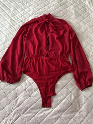 Röd långärmad body med knyt och detaljer - Snygg röd body med långa ärmar, knytning i halsen och svarta dekorativa knappar framtill. Tillverkad i ett mjukt, lätt textilmaterial med resår i midjan för en smickrande passform. Perfekt för dig som vill sticka ut med en trendig look.