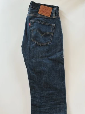 Levi's 501 mörkblå jeans - Klassiska Levi's 501 jeans i mörkblå denim med raka ben och femficksmodell. Snygga kontrastsömmar och ikonisk läderpatch bak i midjan. Jeansen har normal midja och den röda Levi's-taggen på bakfickan. Perfekta för en avslappnad och tidlös stil. De är uppsydda och är då lite kortare än 32 i längd