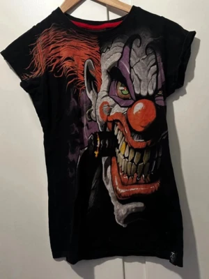 Svart t-shirt med clowntryck - Unik svart t-shirt med stort, färgstarkt tryck av en skrämmande clown på framsidan. T-shirten har korta ärmar och rund halsringning. Perfekt för dig som gillar statement-plagg och vill sticka ut i mängden.