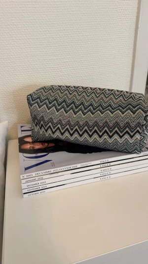 Mönstrad necessär i zigzag-mönster - Säljer en snygg necessär med zigzag-mönster i grönt, svart, vitt och beige. Väskan har en rektangulär form och dragkedja upptill. Perfekt för smink eller småprylar. Materialet känns som tyg och den är mjuk och flexibel.