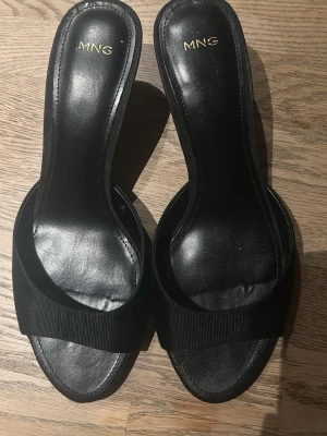 Svarta klackar från mango - Stilrena svarta slip-in pumps från MNG med öppen tå och smal klack. Skorna har bred rem framtill i textil och innersula i konstläder. Perfekta för dig som gillar minimalistisk och elegant design. dem är rätt låga skulle gissa 4 cm