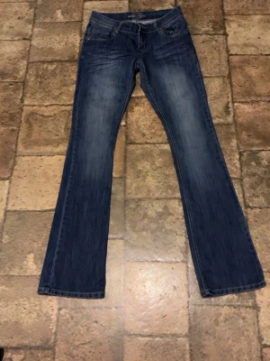 Blå bootcut jeans med slitningar - Säljer ett par blå bootcut jeans med snygga slitningar och lätt tvättad look. Jeansen har klassisk femficksdesign och subtila detaljer på bakfickorna. Perfekta för dig som gillar en avslappnad men trendig stil.