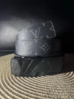 Svart Louis Vuitton monogram skärp - Snyggt svart skärp från Louis Vuitton med klassiskt monogrammönster och stor LV-logga i metall som spänne. Skärpet är gjort i skinn och har en stilren, modern look som passar till många outfits.