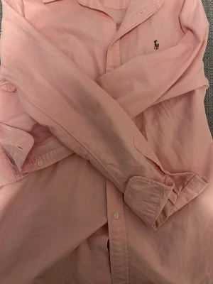 Rosa skjorta från Polo Ralph Lauren - Säljer en klassisk rosa skjorta från Polo Ralph Lauren i custom fit. Skjortan har lång ärm, knappar framtill och den ikoniska broderade loggan på bröstet. Tillverkad i mjuk bomull som känns skön mot huden. Perfekt för dig som gillar stilrena plagg med en preppy vibe.