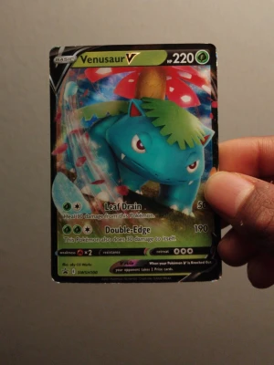 Venusaur V Pokémonkort SWSH100 - Venusaur V är ett samlarkort från Pokémon TCG med holografisk yta. Kortet har färgstark design med Venusaur i blått och grönt, samt detaljer i rött och vitt. Kortet är gjort av laminerad kartong och har attacker som Leaf Drain och Double-Edge. Perfekt för dig som gillar Pokémon och samlarkort.