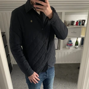 Blå vår jacka Morris  - Säljer en svart quiltad jacka från Morris med klassisk krage och knappstängning framtill. Jackan har ett diskret Morris-emblem på bröstet och är perfekt för dig som gillar stilrena och tidlösa plagg. Passformen är normal och jackan har långa ärmar. Jackan passar perfekt nu när vädret börjar bli lite mildare.