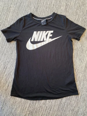 Svart Nike t-shirt med stor logga XS - Snygg svart t-shirt från Nike med stor vit logga på bröstet. Klassisk rund hals och korta ärmar. Tillverkad i mjuk bomull som känns skön mot huden. Perfekt för en sportig och avslappnad stil. Nike Träning T-shirt i storlek XS för dam.   Endast använd vid ett par tillfällen och i bra begagnat skick   Mått Över bröstet: ca 46 cm Längd från axeln och ner: ca 62 cm  52% polyester och 48% modal  Kommer från ett djur och rökfritt hem