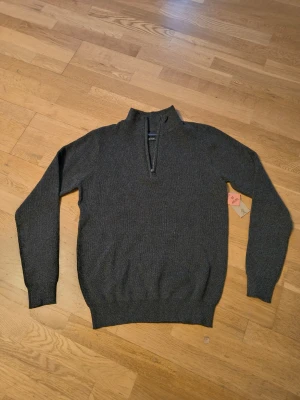 Mörkgrå stickad tröja med halv dragkedja - Mörkgrå stickad tröja med halv dragkedja och hög krage. Tröjan har långa ärmar och ribbade muddar vid ärmslut och nederkant. Perfekt för lager på lager och en stilren look.