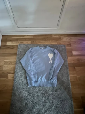 Ljusblå stickad tröja Ami Paris - Ljusblå stickad tröja från Ami Paris med rund halsringning och ribbade muddar. På bröstet finns ett vitt hjärta och bokstaven 'A' som är typiskt för märket. Perfekt för dig som gillar stilrena och trendiga plagg. Använd 2 gånger!