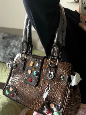 Brun handväska med färgglada stenar - Cool brun handväska i krokodilpräglat konstläder med blanka detaljer och färgglada stenar i olika former. Väskan har dubbla handtag och silverfärgade metalldetaljer. Perfekt för dig som vill sticka ut med en unik accessoar.