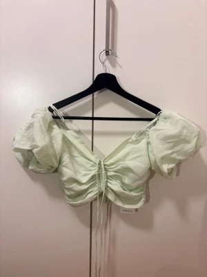 Ljusgrön puffärm topp från Chiquelle - Trendig ljusgrön topp från Chiquelle med puffärmar och rynkade detaljer framtill. Kort modell med snörning i mitten och v-ringad hals. Tillverkad i ett tunt, lätt material som känns luftigt och somrigt.